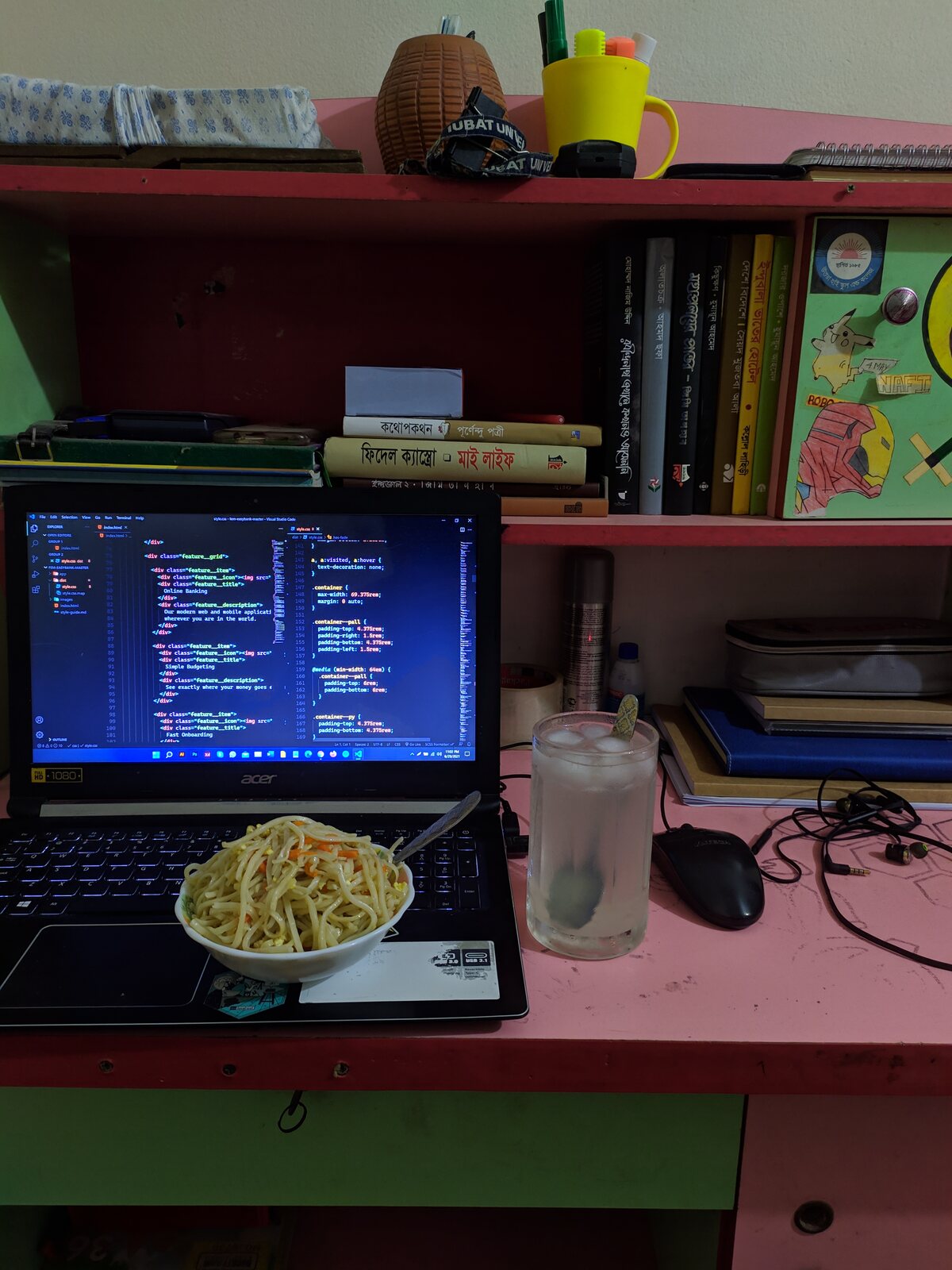 A bowl of noodles and a yellow cup beside a laptop showing code.