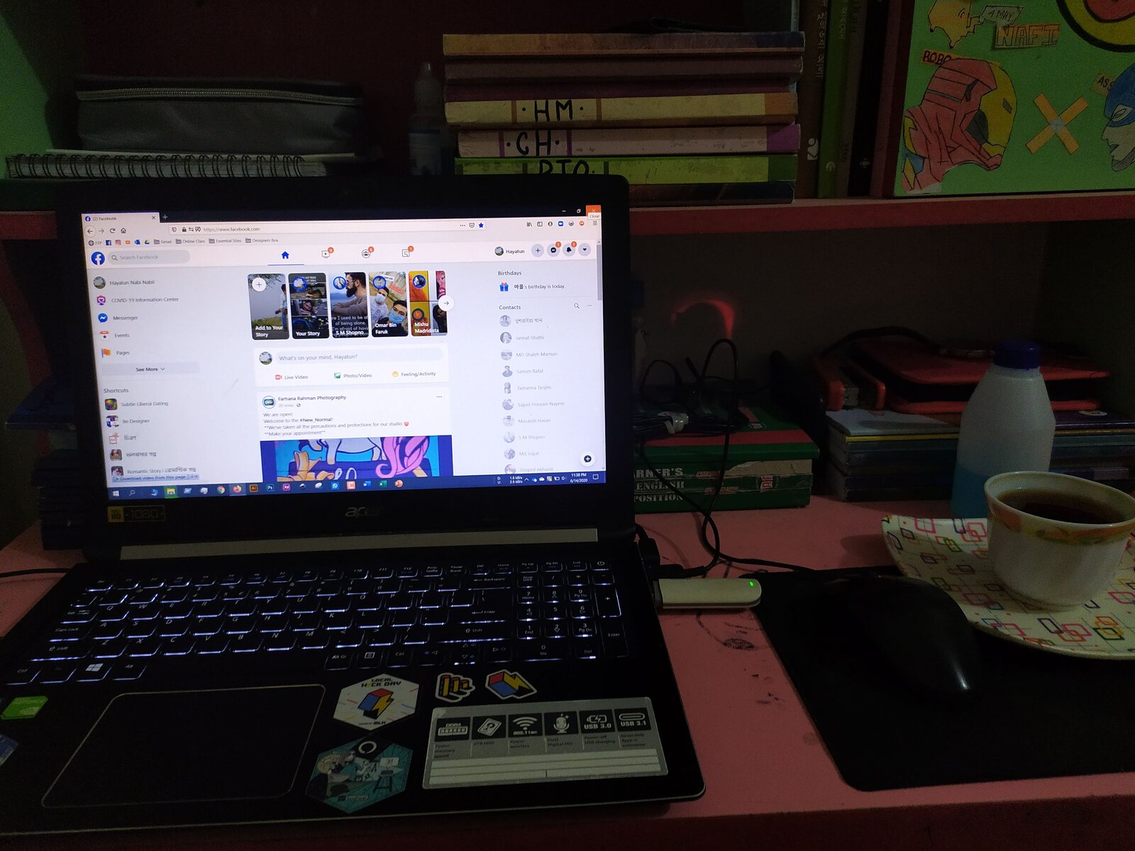 A laptop glowing in a dim room beside a bookshelf and a cup of coffee.