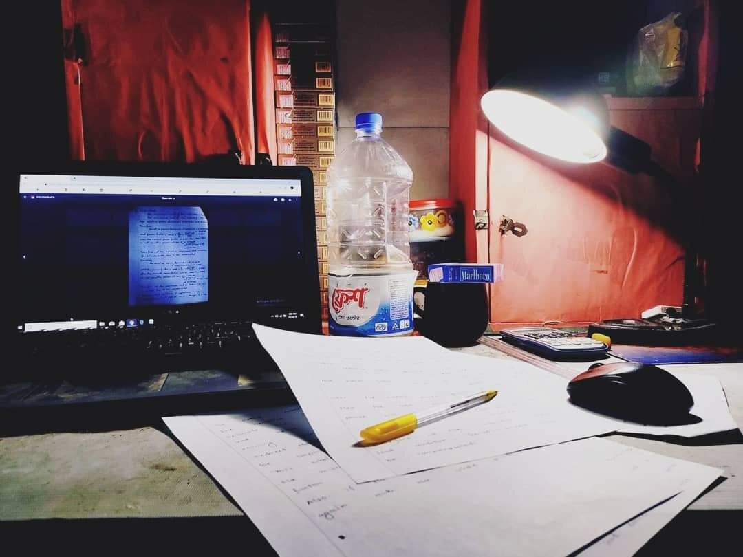 A desk washed in red lamplight with a laptop, papers and a water bottle.